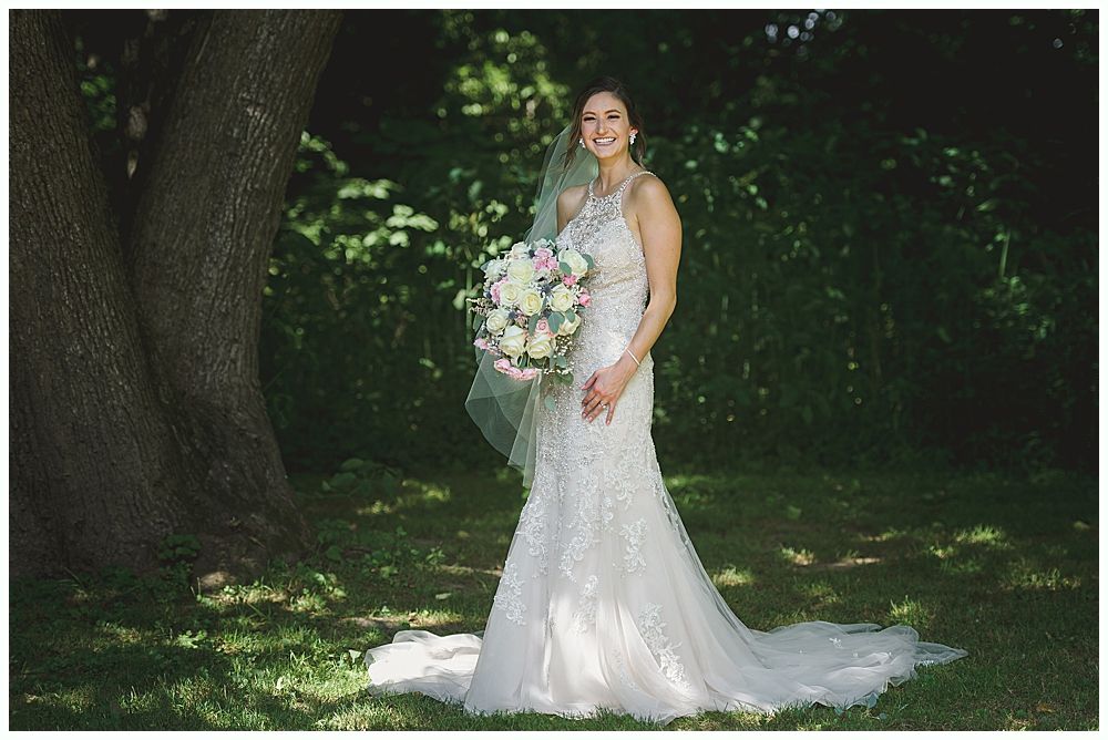 Bride in a beaded wedding dress, holding a bouquet, smiles near a tree in a sunny, green outdoor setting.