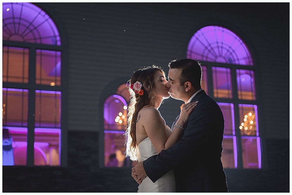 Couple kissing at their wedding, illuminated by purple-lit windows.