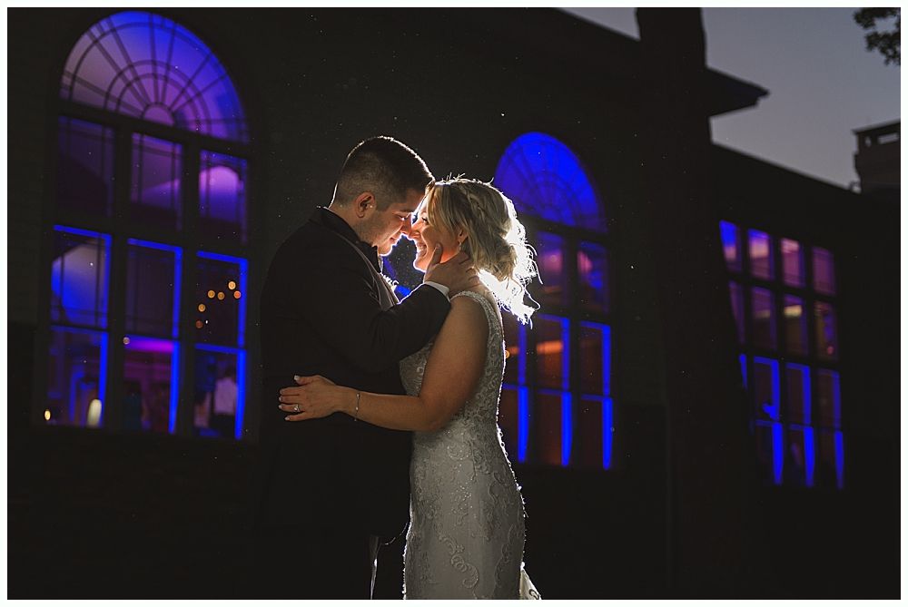 Couple embracing, backlit in front of a building with blue and purple window lights.