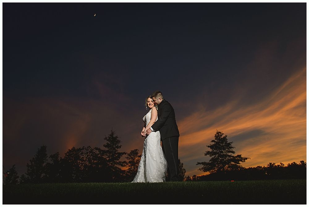 Couple embraces under a vibrant sunset. Bride in white dress, groom in suit on grass, trees in background.