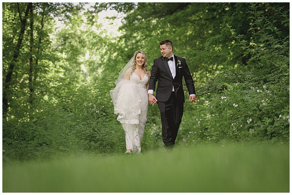 Bride and groom walk hand in hand through a lush green forest, smiling at each other.