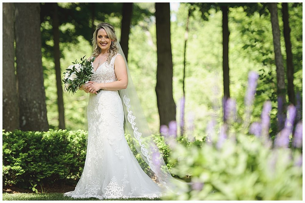 Bride in a white lace wedding dress holding a bouquet, standing in a garden with trees and flowers.
