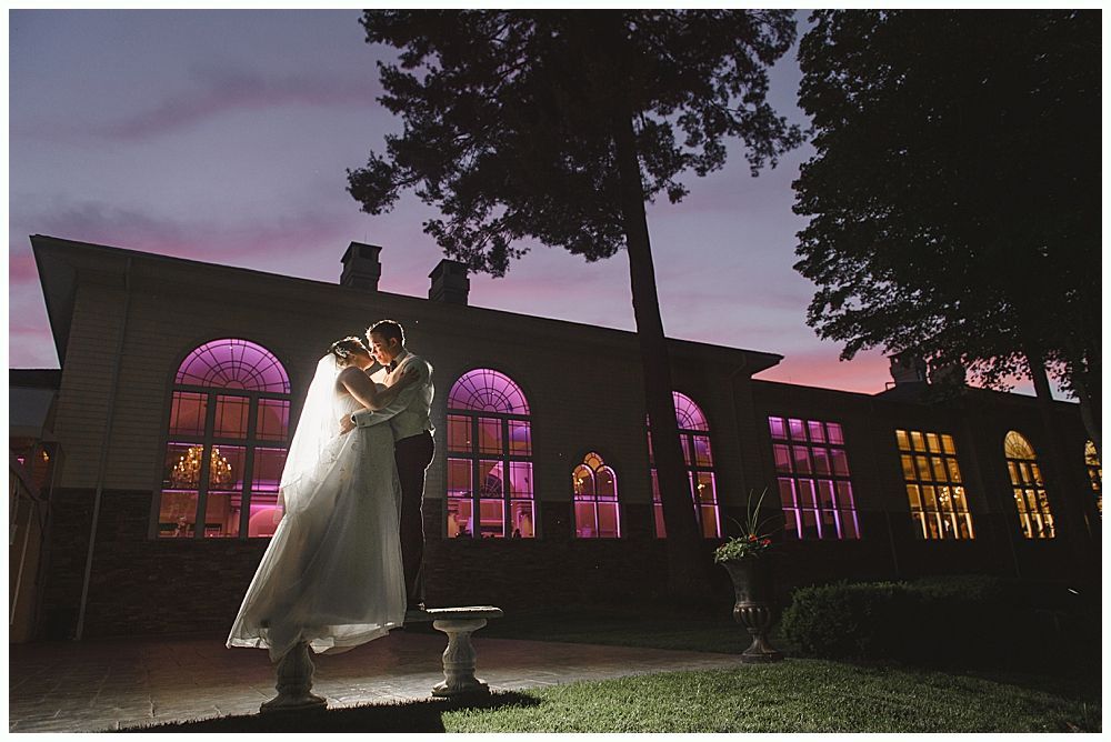 Couple kissing in front of a building with arched windows, lit with colored lights. Sunset sky.