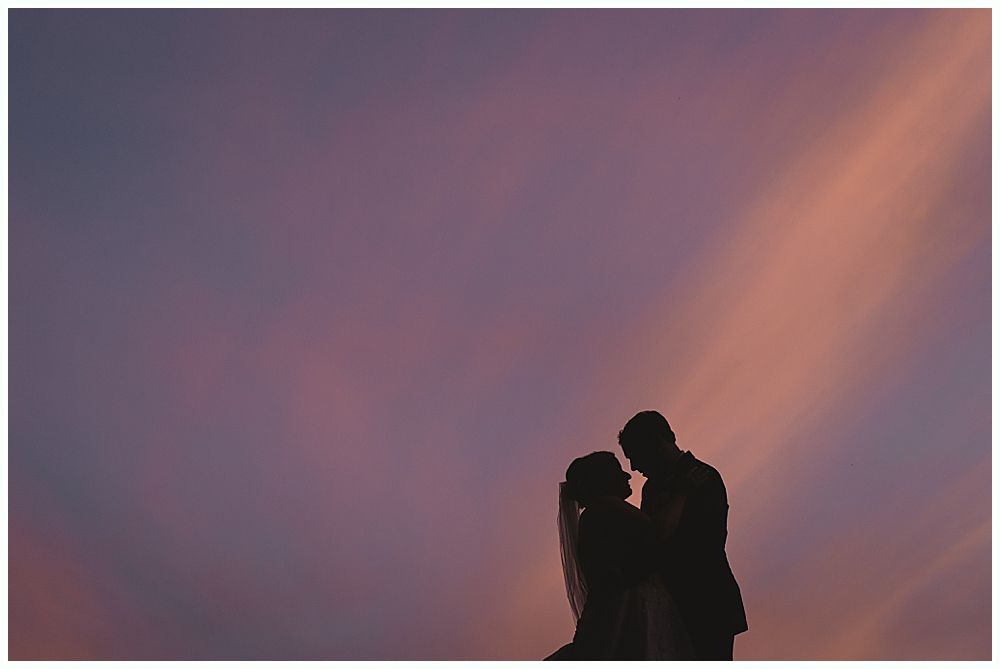 Couple silhouetted against a purple and orange sunset, embracing, likely newlyweds.