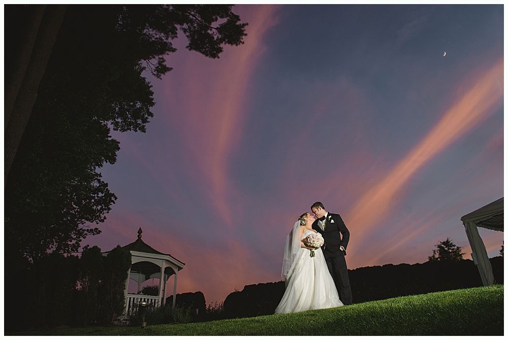 Bride and groom embracing, silhouetted against a colorful sunset sky. Gazebo and trees in background.