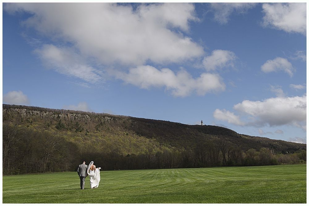 Couple walking on a grassy field; blue sky with puffy white clouds, hill with trees in the background.