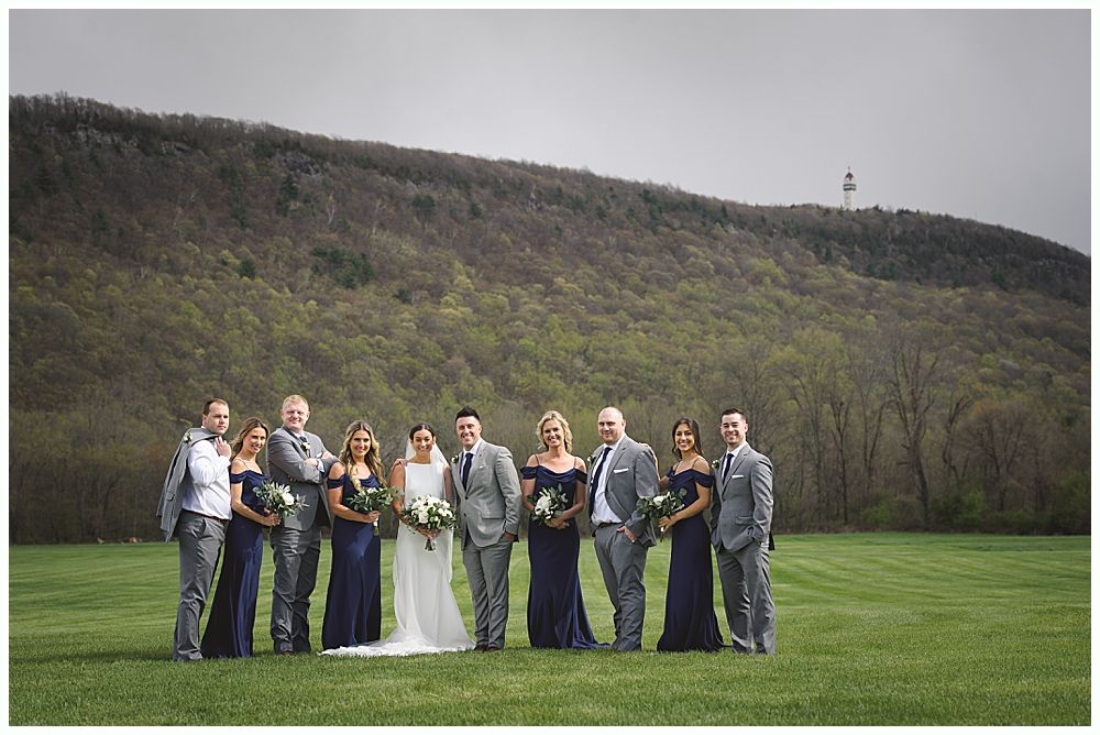 Wedding party posing on a grassy field in front of a wooded mountain. Formal wear: bride in white, bridesmaids in navy, groomsmen in gray.
