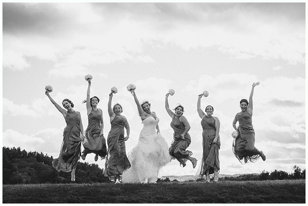 Bride and bridesmaids jumping for joy, holding bouquets, outdoors.