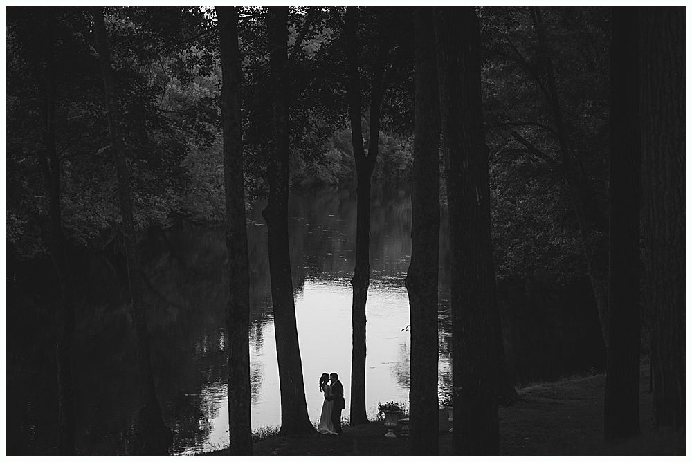 Silhouetted couple by a river, framed by trees. They appear to be embracing. Black and white.