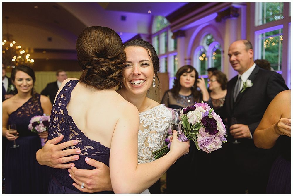 Bride embraces bridesmaid, both smiling at a wedding reception. Purple bouquets, dresses, and decor.