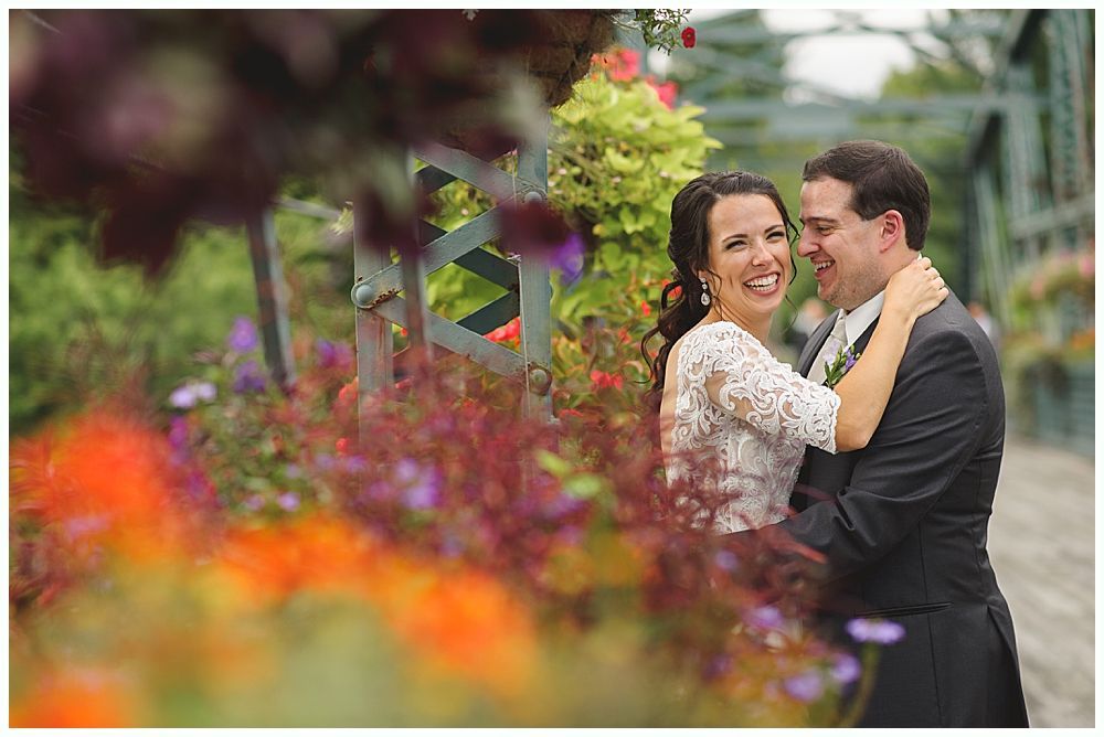 Bride and groom embrace, laughing, near colorful flowers and a bridge.
