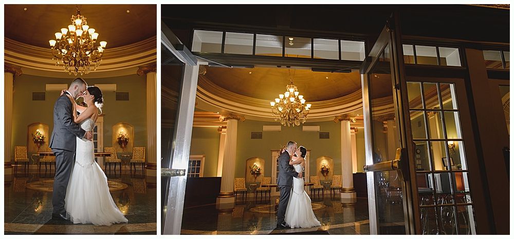 Bride and groom embrace in a ballroom, seen through open doors. They kiss under a chandelier.