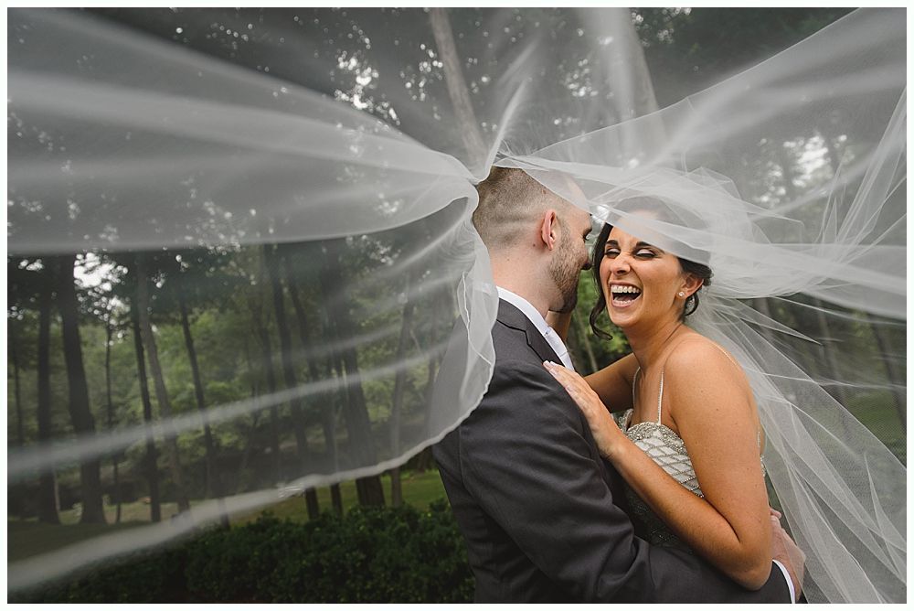 Bride and groom laughing, under a flowing veil; outdoor wedding setting.