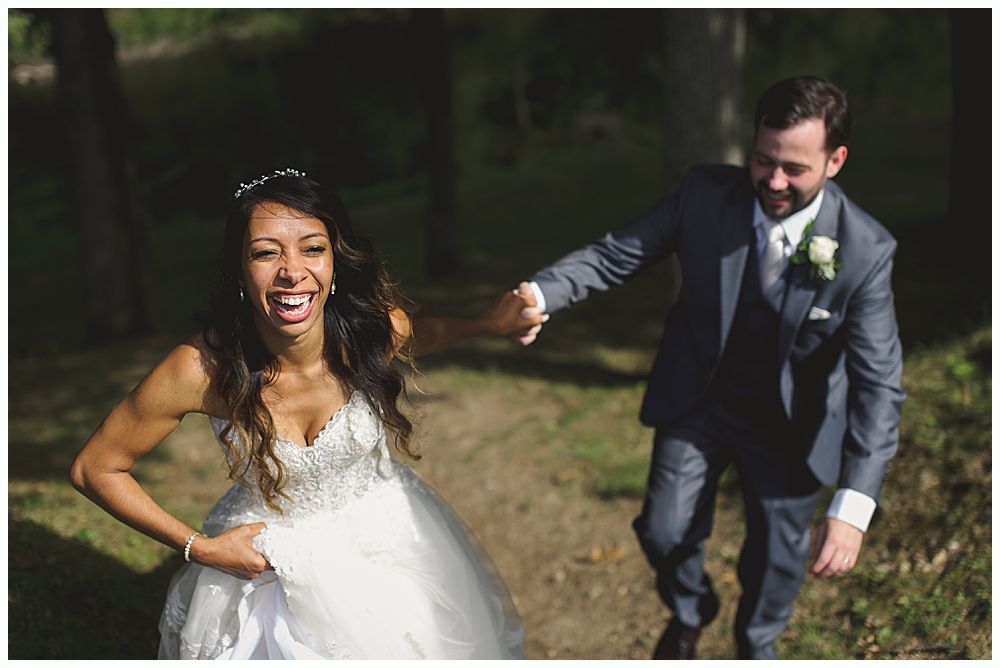 Bride laughing, holding skirt, being led by groom in gray suit on path.