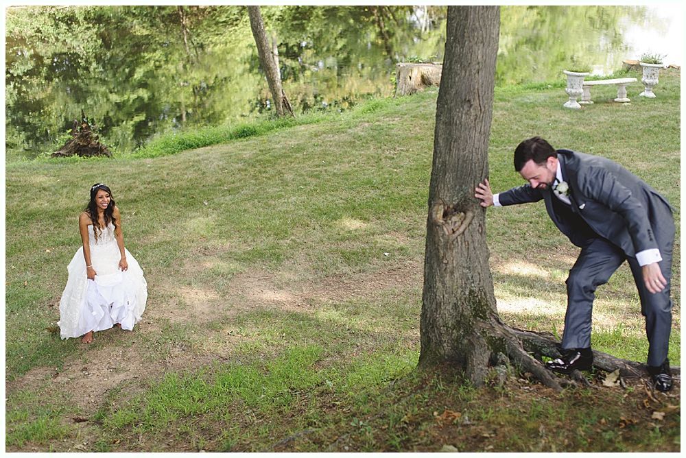 Bride in a white dress sits. Groom in suit peers around a tree in outdoor setting.