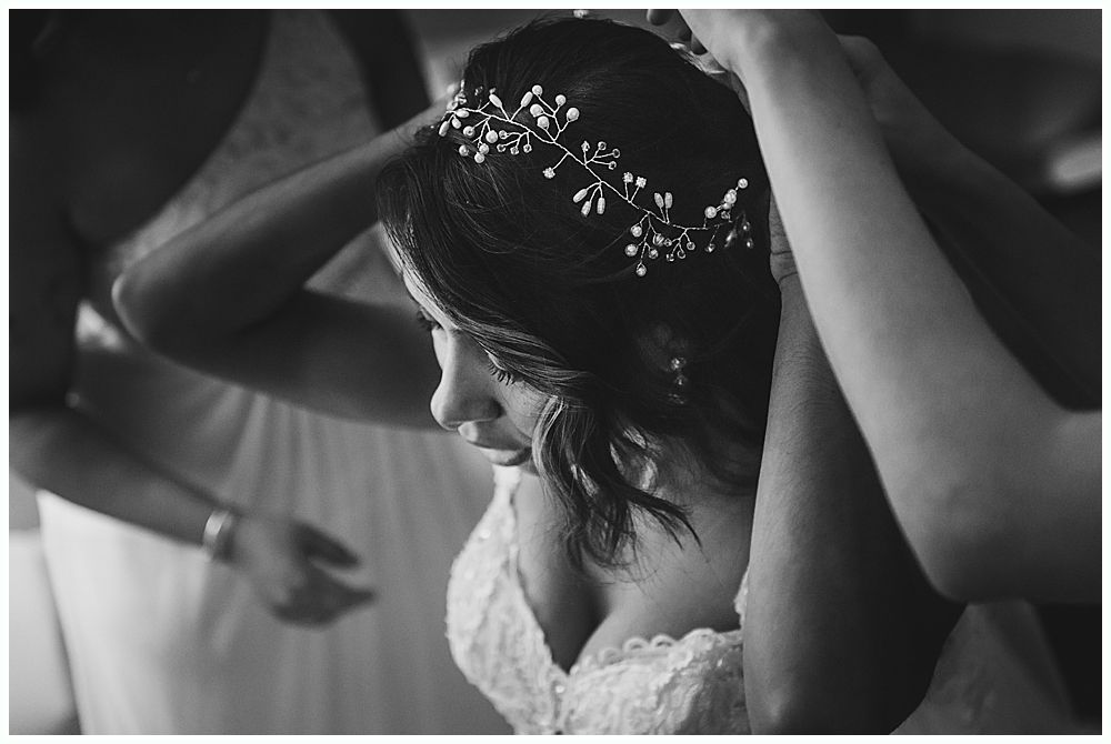 Bride having her hair adorned with a delicate headpiece, bridesmaids assisting in a black and white photo.