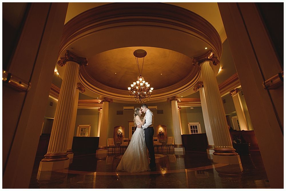 A couple embracing under a domed ceiling, inside a grand building with pillars and chandelier.