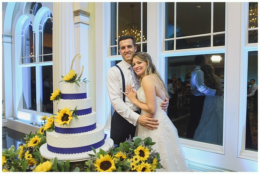 Newlyweds pose by wedding cake decorated with sunflowers, beside a window overlooking a reception.