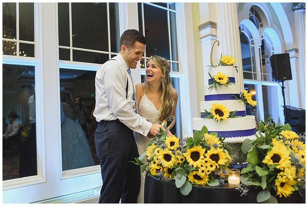 Couple cutting wedding cake, sunflowers, laughing. White tiered cake with navy ribbon. Large window background.