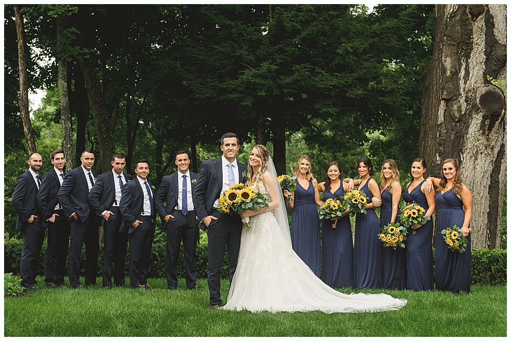 Wedding party poses on a lawn. Bride and groom in center, flanked by groomsmen and bridesmaids in navy with sunflowers.
