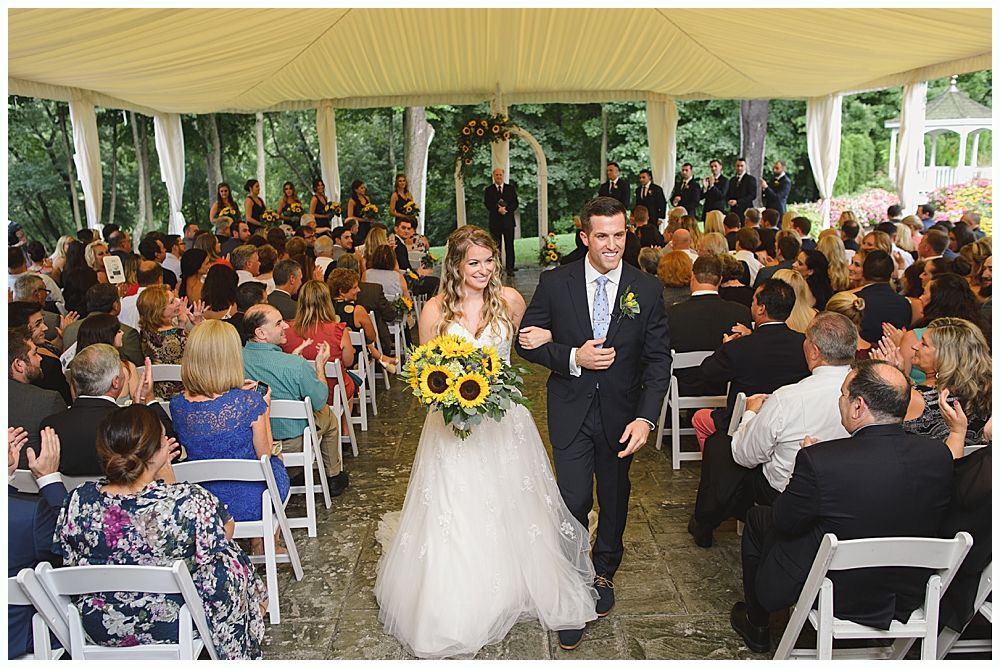 Bride and groom walk down aisle, sunflowers in bouquet, guests seated in tent.