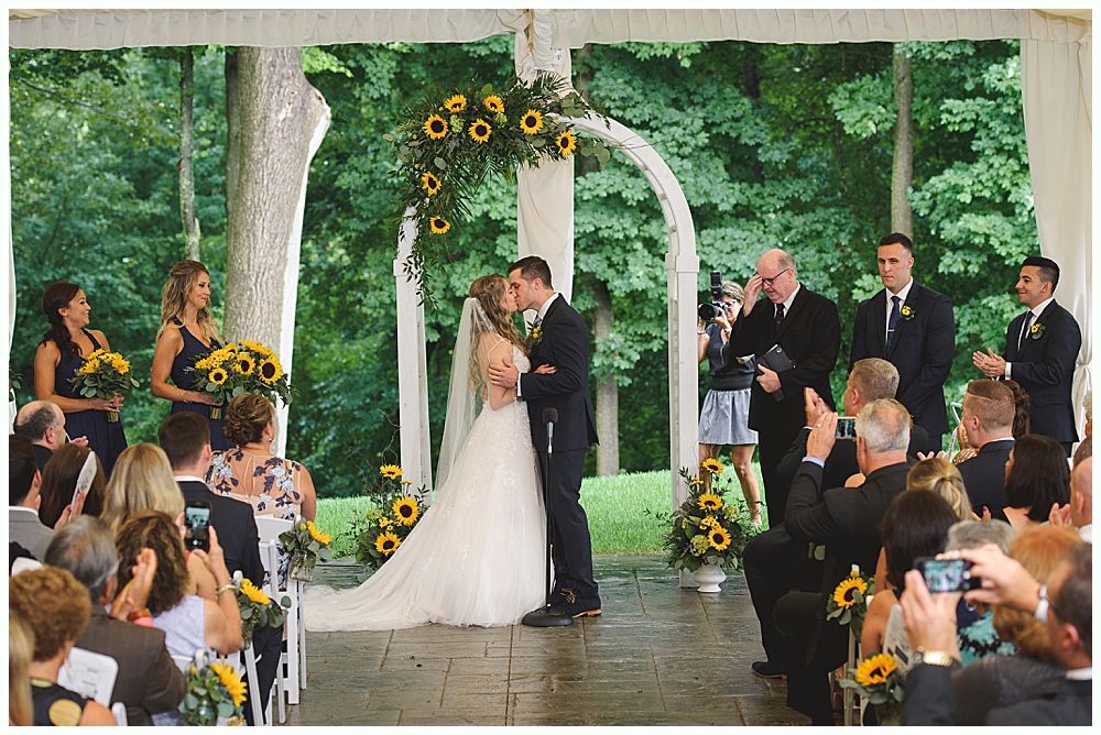 Wedding ceremony: couple kissing under floral arch, surrounded by guests and wedding party.