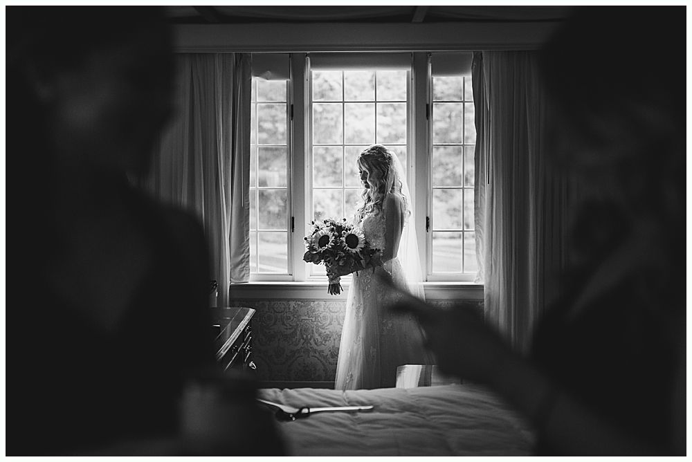 Bride in wedding dress holds flowers by a window; bridesmaids in foreground.