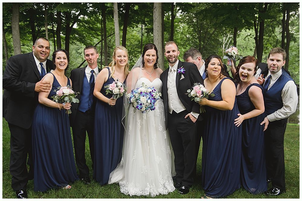 Wedding party: Bride in white dress surrounded by bridesmaids in blue, groomsmen in black, outdoors.