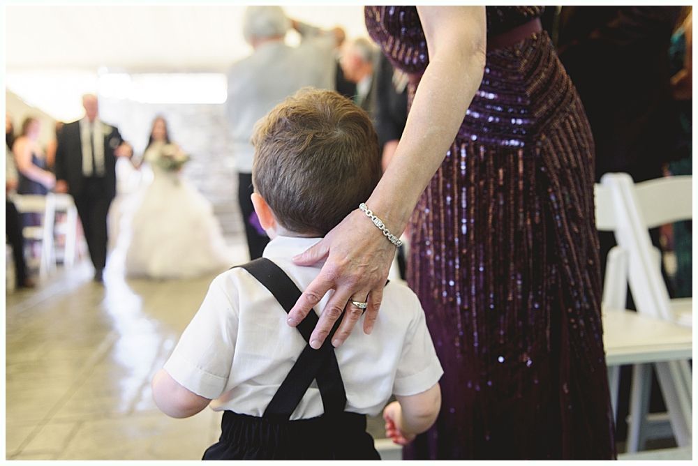 Boy in suspenders watched a bride walk down the aisle, hand on his back.