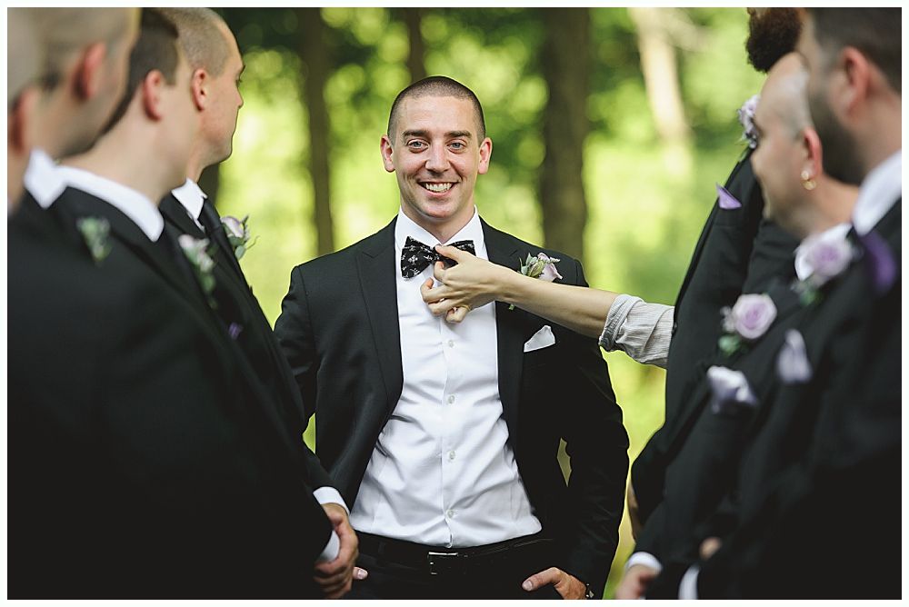 Groom smiles, adjusting his bow tie, surrounded by groomsmen in black suits, outdoors.