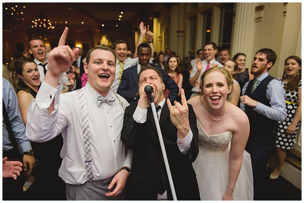 Wedding reception: joyful group dancing, bride and groom smiling with band member, others cheering, formal attire.