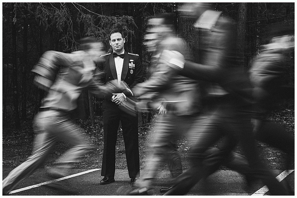 Man in suit standing still as blurred figures in uniform run past him. Outdoor setting.