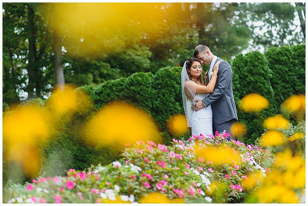 Bride and groom embrace in a garden with colorful flowers. They are smiling, with a lush green backdrop.