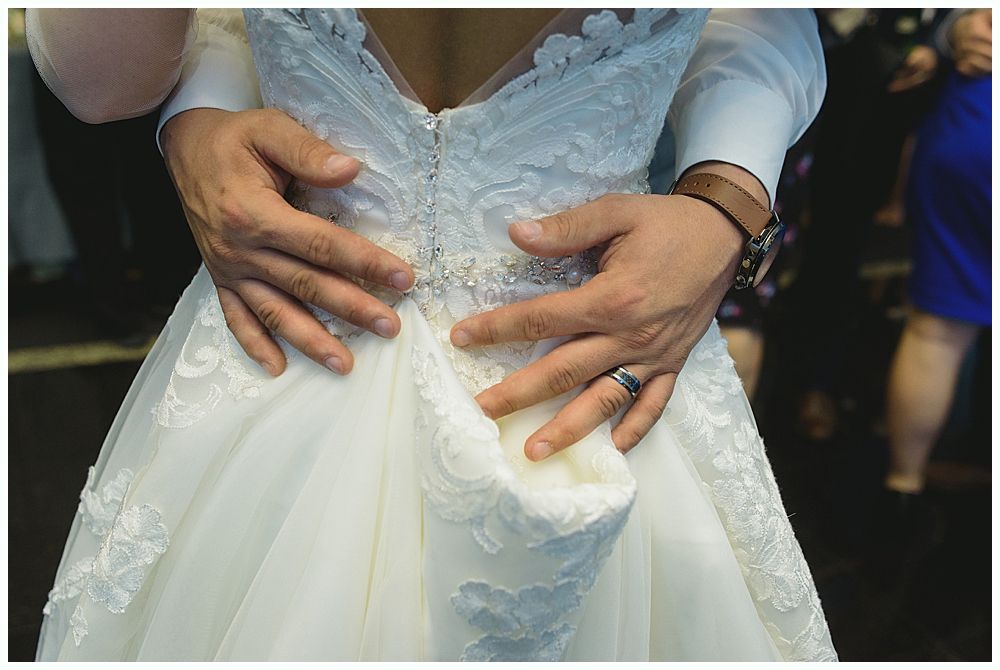 Hands on the back of a wedding dress, with the groom's hands clasped around the bride.