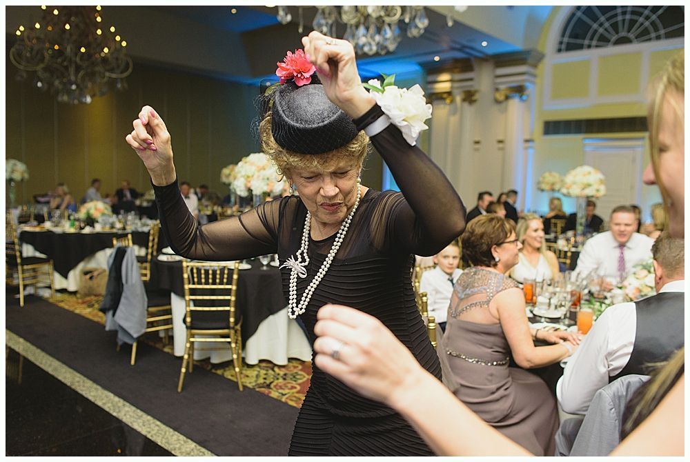 Woman dancing at a wedding reception; wearing a black dress and hat; many guests are seated at tables.