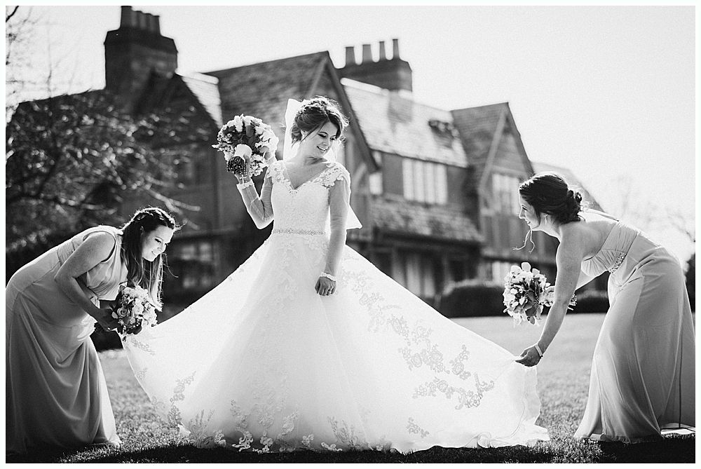 Bride with bridesmaids holding train of her wedding dress in front of a large house. Black and white.