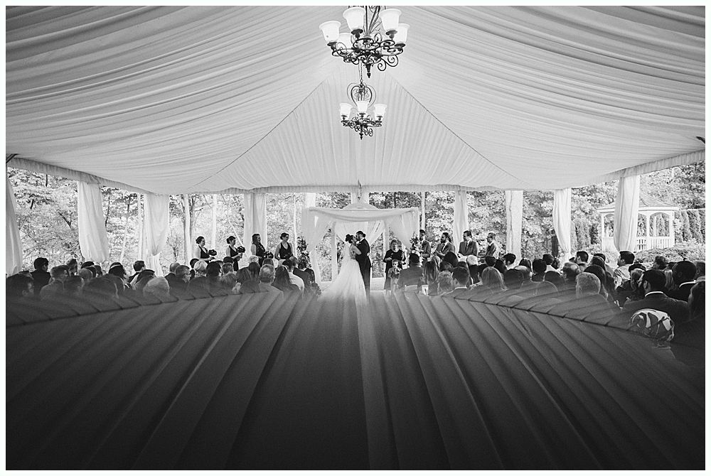 Wedding ceremony under a tent with couple at the altar; guests seated on either side, chandeliers overhead.