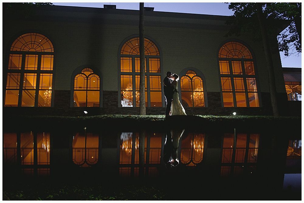 Couple embracing in front of a building with lit windows reflecting in water at night.
