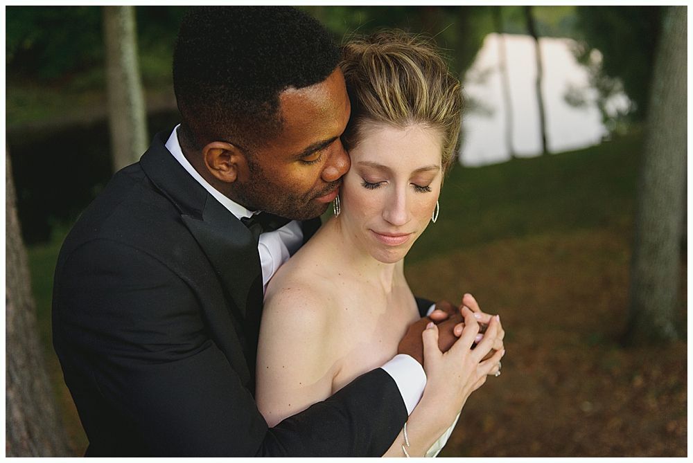 Man in tuxedo kisses woman's cheek; they embrace, outdoors.