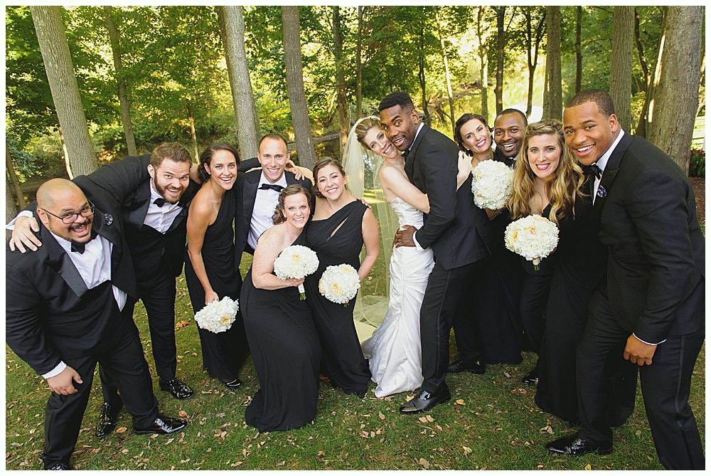 Wedding party smiles for photo outdoors. Bride, groom, bridesmaids, groomsmen, wearing black attire, holding bouquets.