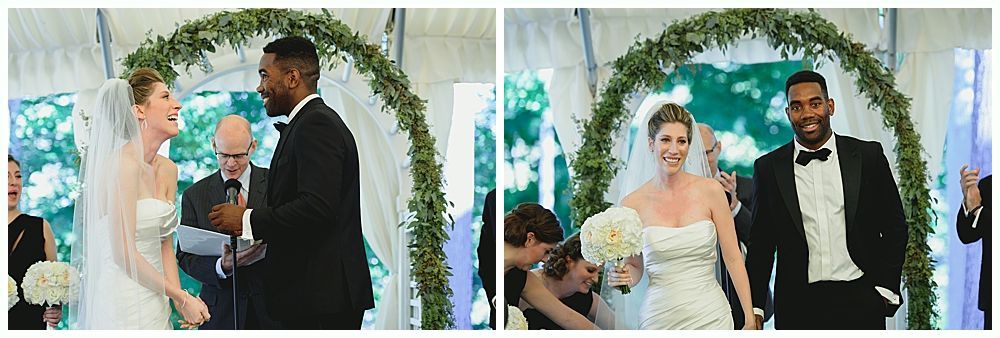 Bride and groom at wedding ceremony under floral arch. They both smile, outdoors.