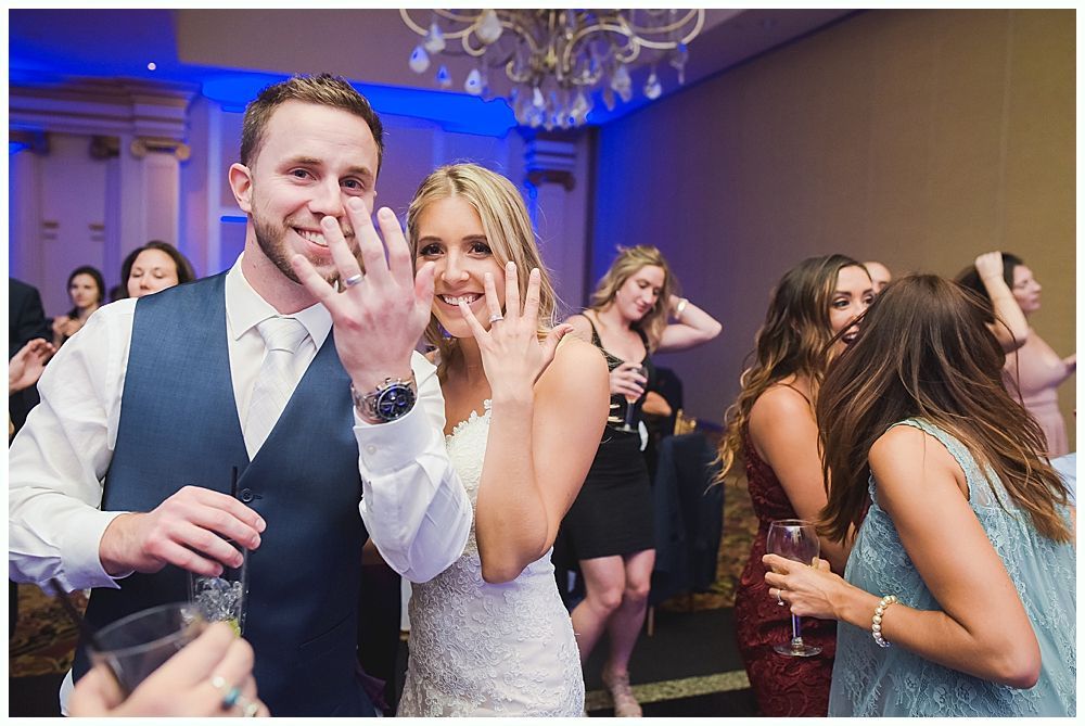 Newlyweds show off rings at a wedding reception. People dance and celebrate in a brightly lit room.