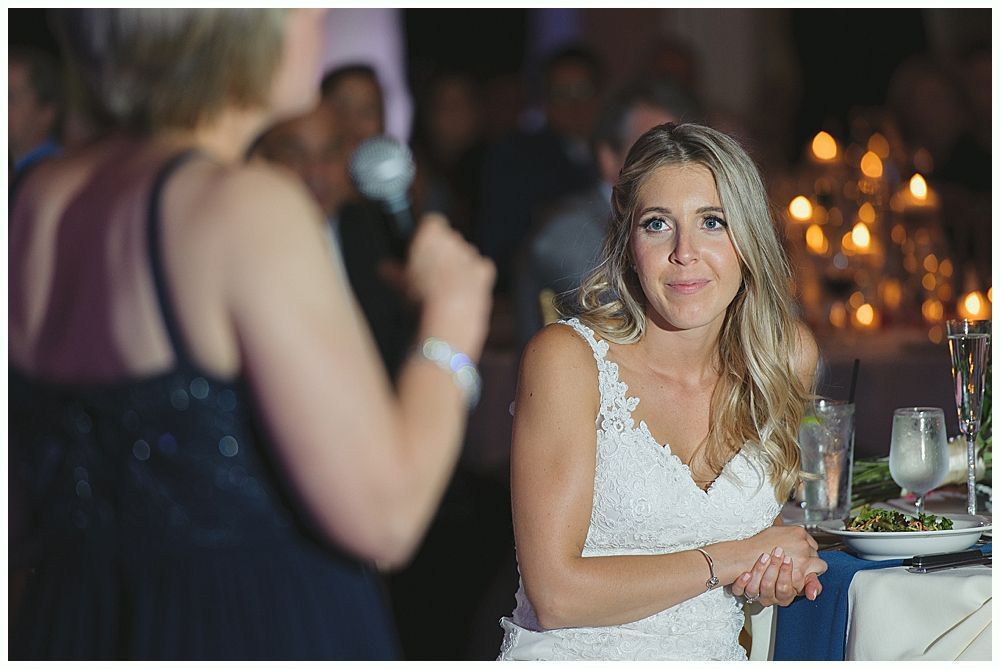 Woman in white lace dress smiles, listening to a speech at a candlelit event.