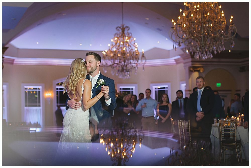 Couple dances at wedding reception; ornate chandelier hangs above them, guests watch.