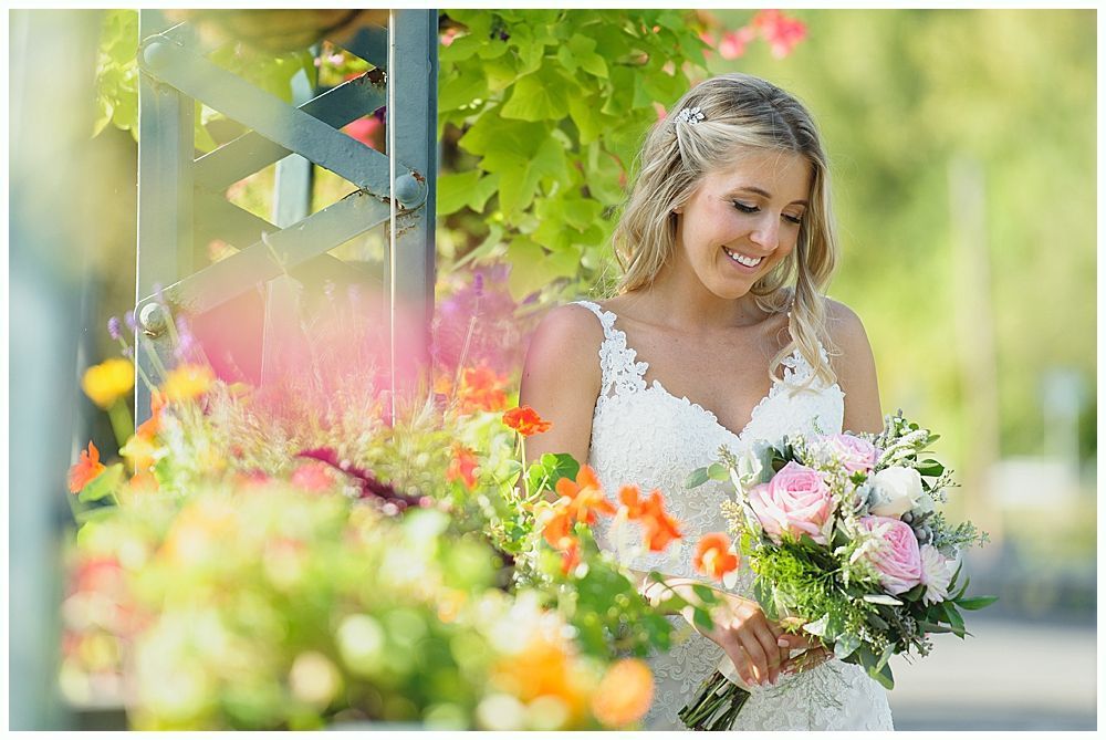 Bride in white lace dress, holding a bouquet, smiles near colorful flowers and greenery.