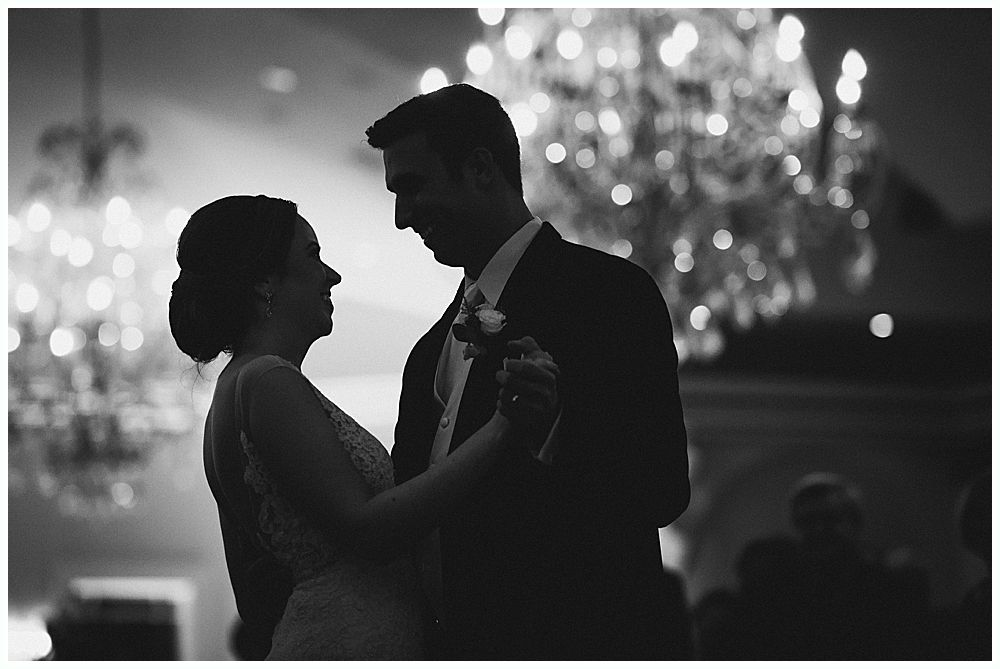 Couple dancing, silhouetted. Bride in wedding dress, groom in suit, under chandeliers.