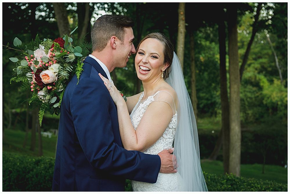 Bride laughs as groom whispers in her ear outdoors with floral bouquet.