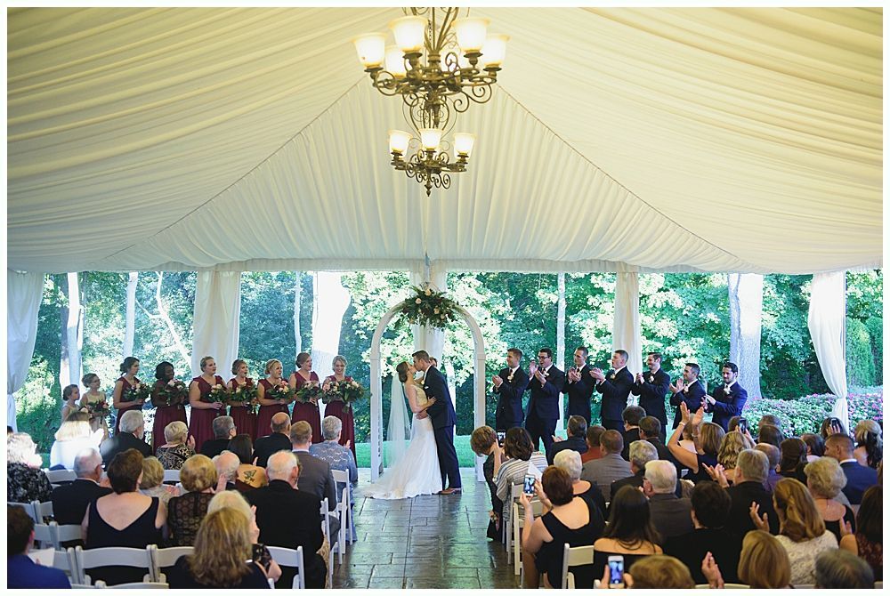 Wedding ceremony under a white tent. Bride and groom kiss, bridal party stands behind them, guests in chairs.