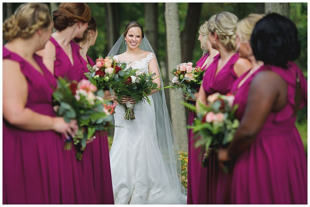 Bride in white dress smiles, surrounded by bridesmaids in magenta gowns, holding bouquets.