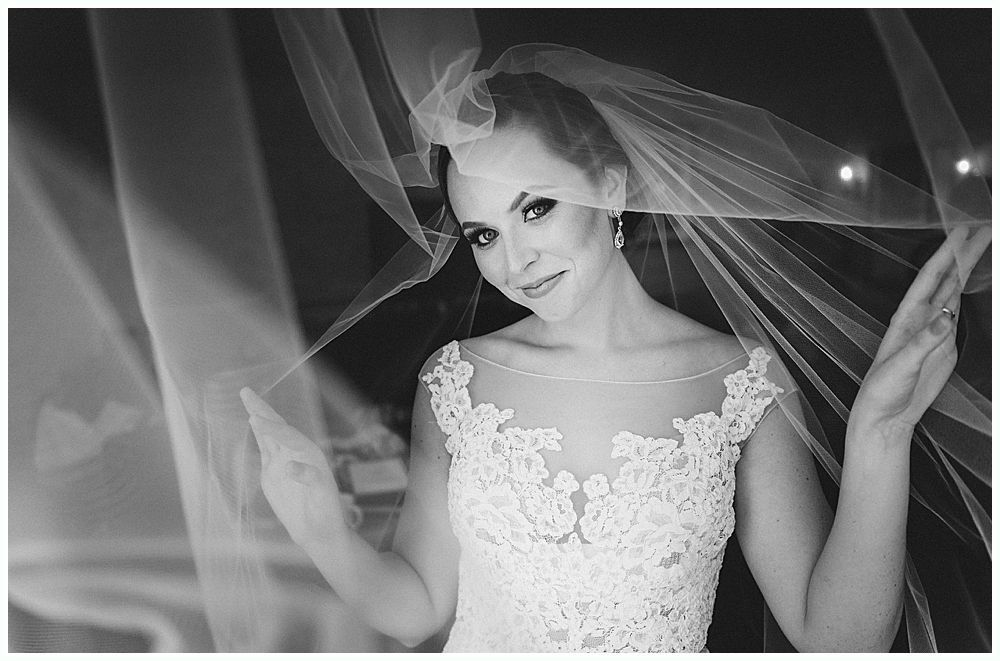 Bride in a lace wedding dress, veil framing her face, holding up the veil, smiling. Black and white.